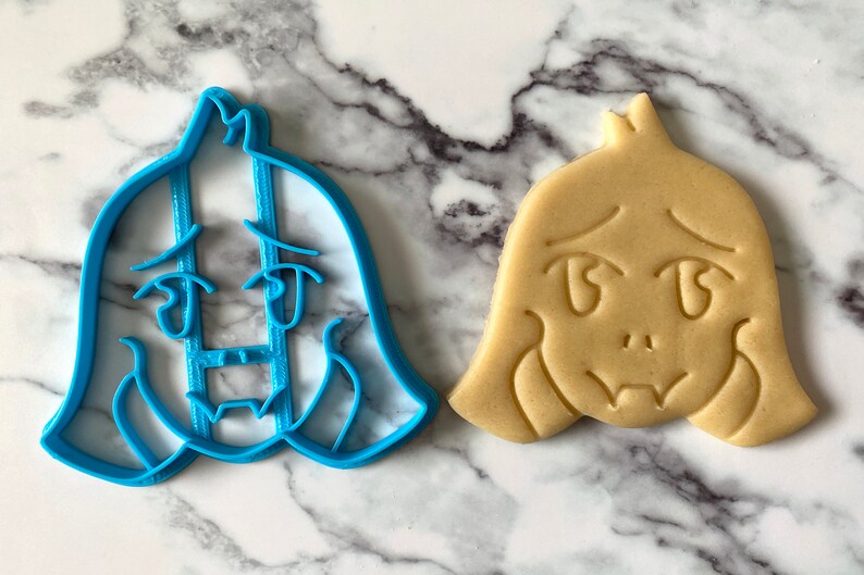 May include: A blue plastic cookie cutter shaped like a cartoon character with a sad expression, next to a light brown cookie with the same design.