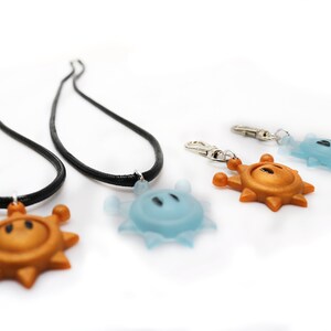 May include: Two black necklaces with gold and blue sun-shaped pendants. A separate gold sun-shaped pendant is attached to a silver key chain.