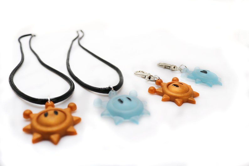 May include: Two black necklaces with gold and blue sun-shaped pendants. A separate gold sun-shaped pendant is attached to a silver key chain.