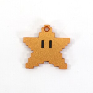 May include: A gold-colored star-shaped charm with a pixelated design and two black eyes. The charm has a small hole at the top for hanging.
