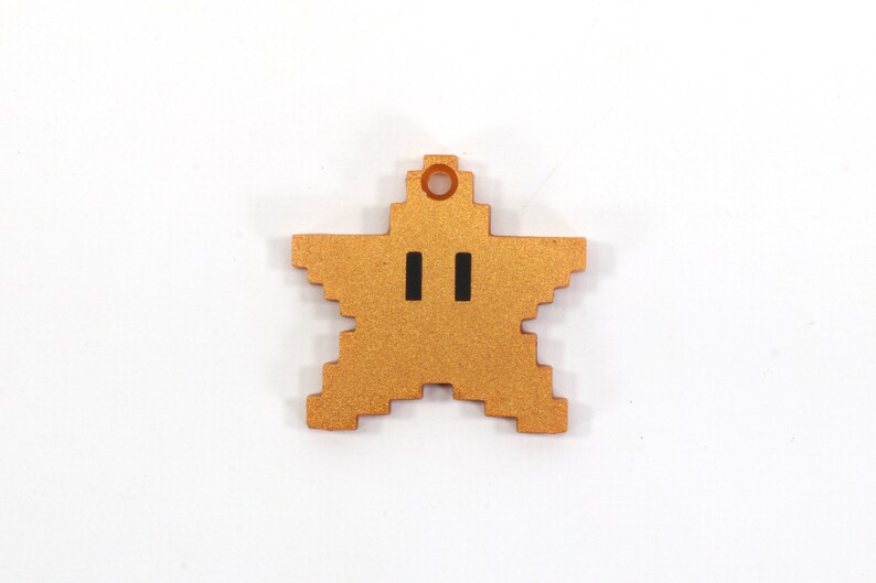 May include: A gold-colored star-shaped charm with a pixelated design and two black eyes. The charm has a small hole at the top for hanging.