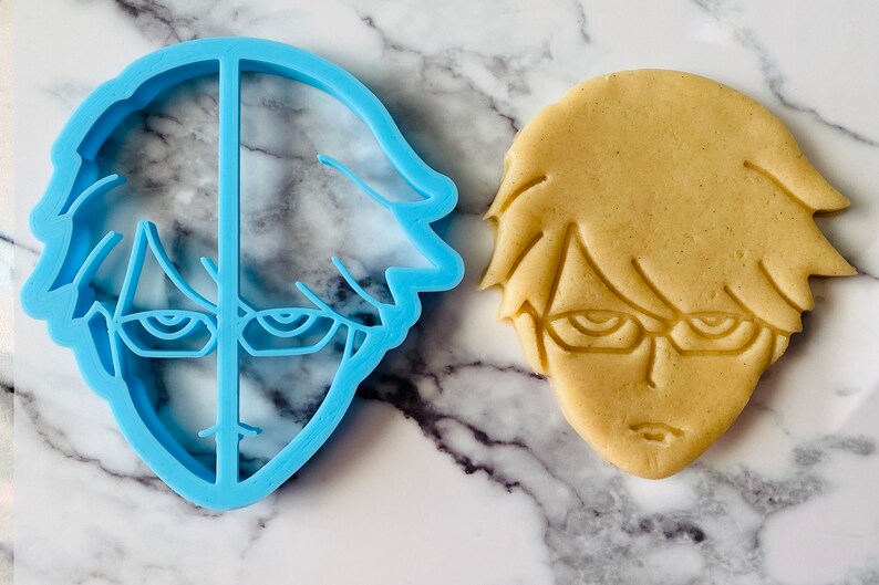 May include: A blue plastic cookie cutter and a light brown cookie in the shape of a cartoon character with glasses and short hair.