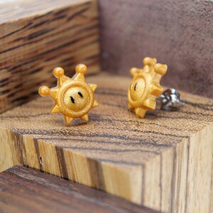 May include: A pair of gold-coloured earrings shaped like a sun with a face. The earrings are on a wooden surface.