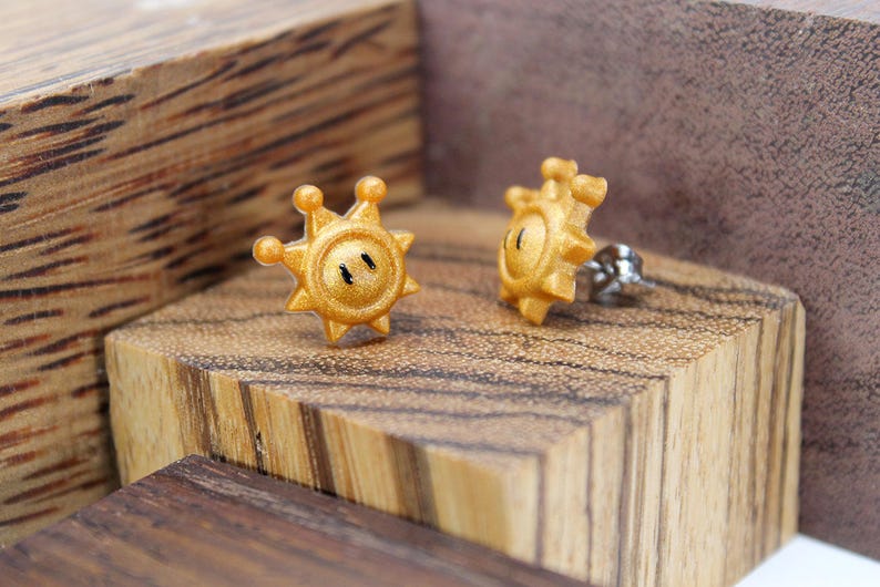 May include: A pair of gold-coloured earrings shaped like a sun with a face. The earrings are on a wooden surface.