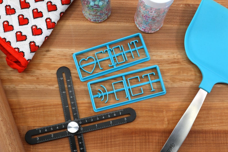 May include: Two blue plastic cookie cutters on a wooden surface. The cutters are shaped like the words "FIGHT" and "ACT".