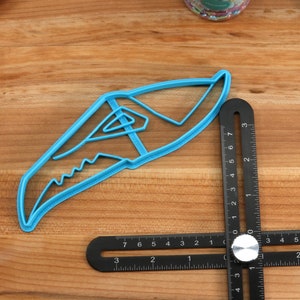 May include: A blue plastic cookie cutter shaped like a shark fin. The cutter is on a wooden surface next to a black ruler.