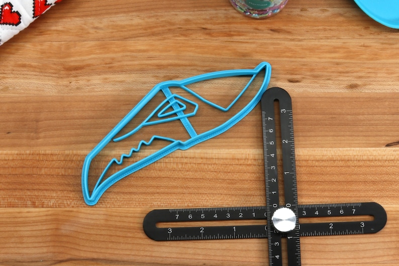 May include: A blue plastic cookie cutter shaped like a shark fin. The cutter is on a wooden surface next to a black ruler.