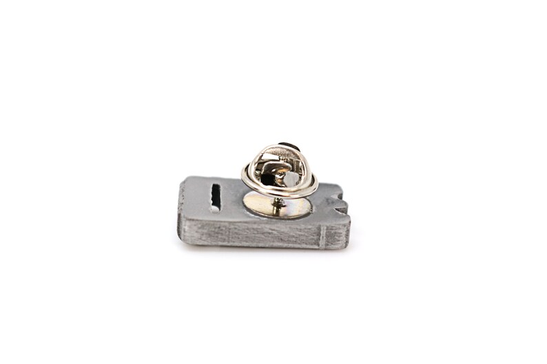 May include: A silver-toned metal pin shaped like a cassette tape with a silver-toned metal pin back.