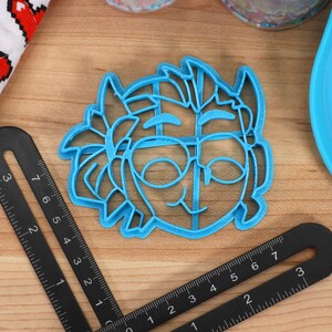 May include: A blue plastic cookie cutter shaped like a cartoon character with glasses and a smile.
