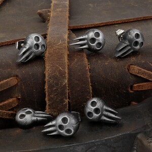 Peut inclure: Six boucles d'oreilles en argent en forme de cr&acirc;ne avec trois trous dans chaque cr&acirc;ne et un motif d'aile sur le c&ocirc;t&eacute;. Les boucles d'oreilles sont pr&eacute;sent&eacute;es sur une surface en cuir marron.