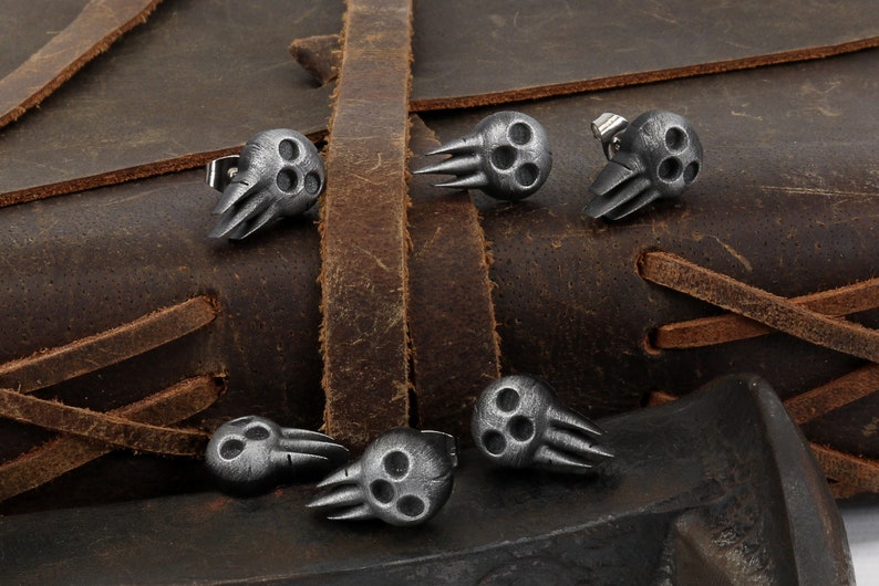 Peut inclure: Six boucles d'oreilles en argent en forme de cr&acirc;ne avec trois trous dans chaque cr&acirc;ne et un motif d'aile sur le c&ocirc;t&eacute;. Les boucles d'oreilles sont pr&eacute;sent&eacute;es sur une surface en cuir marron.