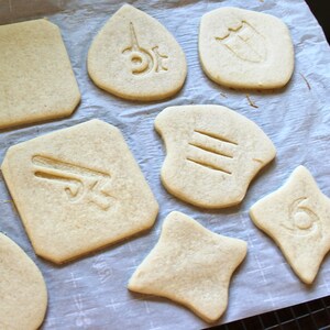 May include: A close-up of a baking sheet with various cookie shapes, each with a different design. The cookies are light brown and have a slightly textured surface. The designs include a sword, a shield, a star, and a number three.