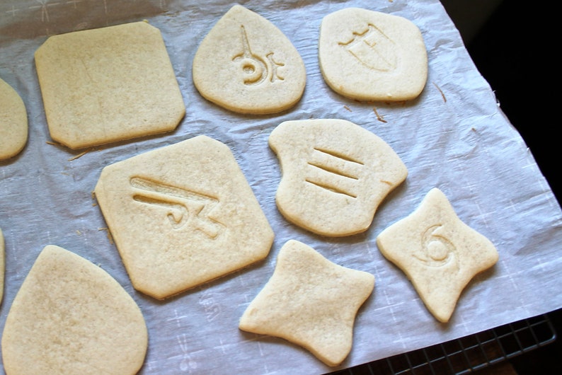 May include: A close-up of a baking sheet with various cookie shapes, each with a different design. The cookies are light brown and have a slightly textured surface. The designs include a sword, a shield, a star, and a number three.