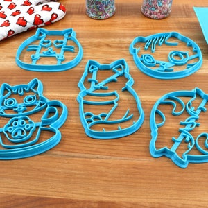 May include: Set of five blue plastic cat-shaped cookie cutters. The cutters feature different cat poses, including a cat in a cup, a cat sitting, and a cat curled up.