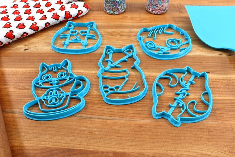 May include: Set of five blue plastic cat-shaped cookie cutters. The cutters feature different cat poses, including a cat in a cup, a cat sitting, and a cat curled up.