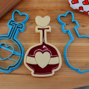 May include: Three blue plastic cookie cutters shaped like a bottle with a heart on top. The middle cutter has a cookie inside with red icing and a white heart.