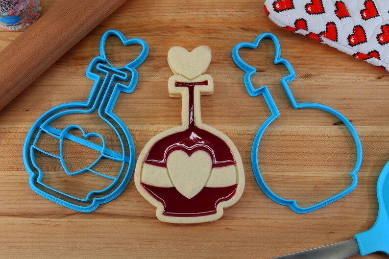 May include: Three blue plastic cookie cutters shaped like a bottle with a heart on top. The middle cutter has a cookie inside with red icing and a white heart.