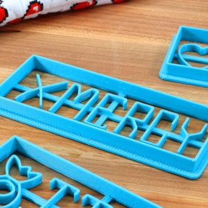 May include: Three blue plastic cookie cutters, one shaped like a heart, one shaped like a Christmas tree, and one shaped like the word "Merry".