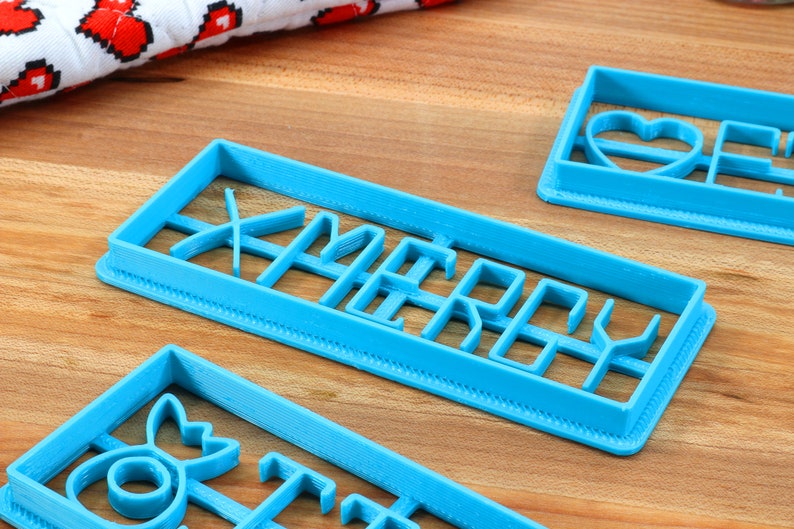 May include: Three blue plastic cookie cutters, one shaped like a heart, one shaped like a Christmas tree, and one shaped like the word "Merry".