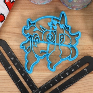 May include: A blue plastic cookie cutter shaped like a cartoon character with hearts and horns. The cutter is on a wooden surface with a ruler.