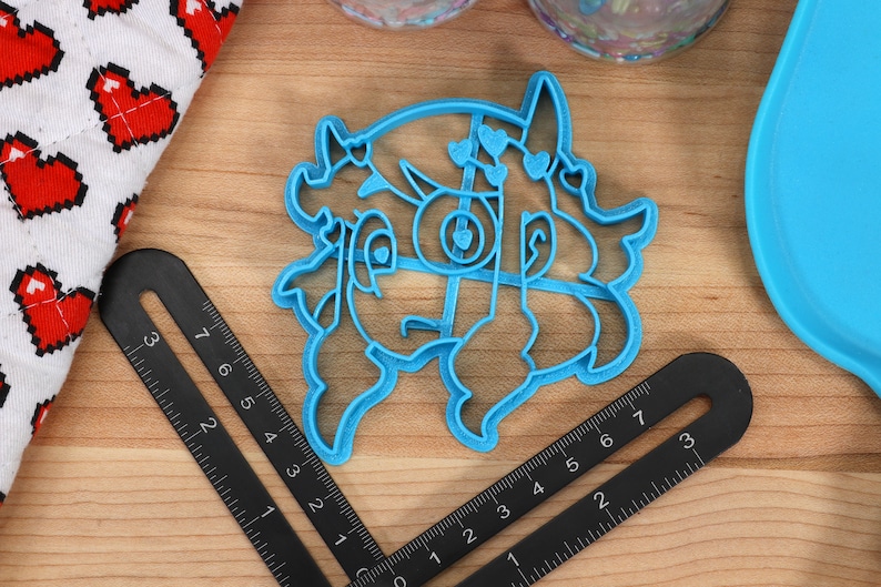 May include: A blue plastic cookie cutter shaped like a cartoon character with hearts and horns. The cutter is on a wooden surface with a ruler.