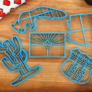May include: A set of five blue cookie cutters shaped like a cactus, a state of Arizona, a road sign with "Arizona" written on it, a silhouette of a bear, and the Arizona state flag.