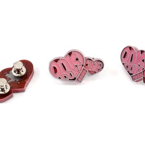 May include: Three pink heart-shaped pins with the text "DOK DOK PARTY CLUB" in white. The pins have a silver metal finish and a safety pin closure.