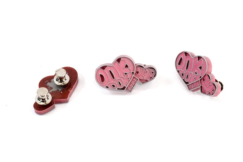 May include: Three pink heart-shaped pins with the text "DOK DOK PARTY CLUB" in white. The pins have a silver metal finish and a safety pin closure.