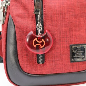 May include: Red crossbody bag with a black zipper and a red and black charm attached to the zipper pull. The charm is shaped like a crescent moon with a symbol in the center. The bag has a black leather trim and a small black label with the text "Where is the heart to receive good where it is" on it.