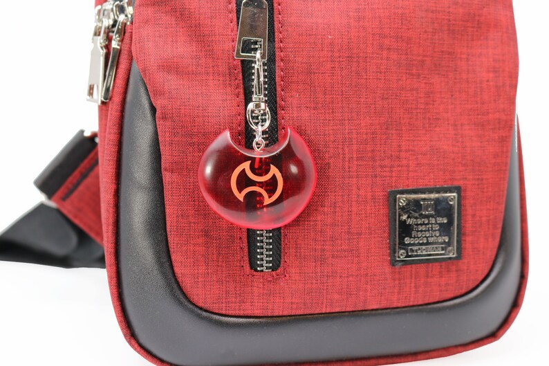 May include: Red crossbody bag with a black zipper and a red and black charm attached to the zipper pull. The charm is shaped like a crescent moon with a symbol in the center. The bag has a black leather trim and a small black label with the text "Where is the heart to receive good where it is" on it.