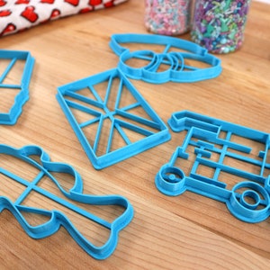 May include: Five blue plastic cookie cutters on a wooden surface. The cutters are shaped like a fish, a truck, a flag, a bee, and a rectangle.