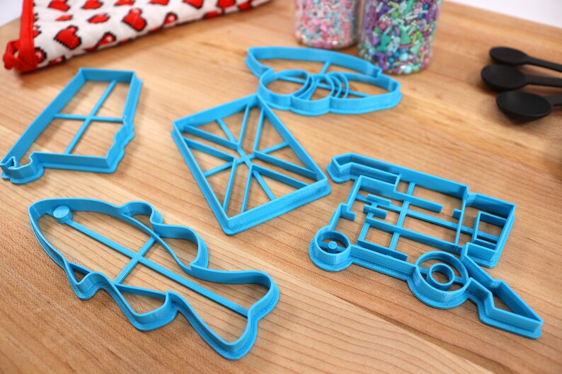 May include: Five blue plastic cookie cutters on a wooden surface. The cutters are shaped like a fish, a truck, a flag, a bee, and a rectangle.