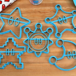 May include: Five blue plastic cookie cutters shaped like stars, a sun, a moon, and a pixelated character. The cutters are on a wooden surface.