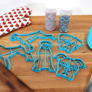 May include: A set of blue plastic dog-shaped cookie cutters, a wooden rolling pin, a blue spatula, and two jars of colorful sprinkles on a wooden cutting board.