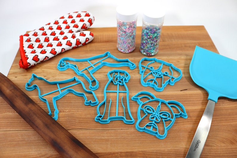 May include: A set of blue plastic dog-shaped cookie cutters, a wooden rolling pin, a blue spatula, and two jars of colorful sprinkles on a wooden cutting board.