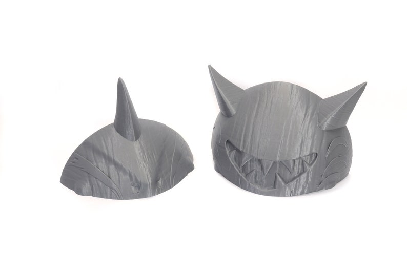 May include: Two gray 3D printed masks, one with a single horn and the other with two horns and a toothy grin.