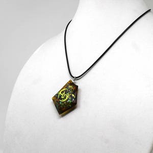 May include: A black cord necklace with a brown, green, and gold pendant featuring a yellow symbol. The pendant is shaped like a diamond.