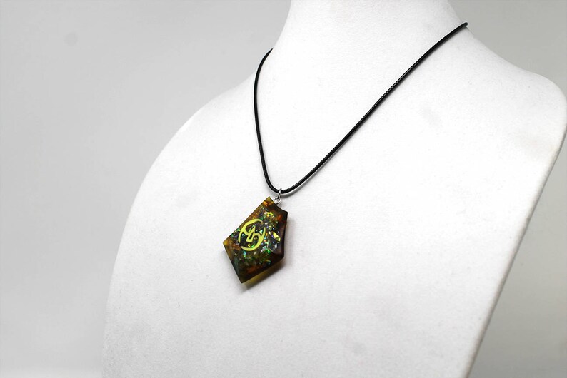 May include: A black cord necklace with a brown, green, and gold pendant featuring a yellow symbol. The pendant is shaped like a diamond.