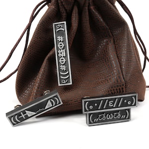 May include: A brown leather pouch with a drawstring closure and four silver metal tags with various symbols and text, including "((#Ф益# 6 &Phi;# 19)", "(..//E//..)", and "(.. డబ్బా ..)".