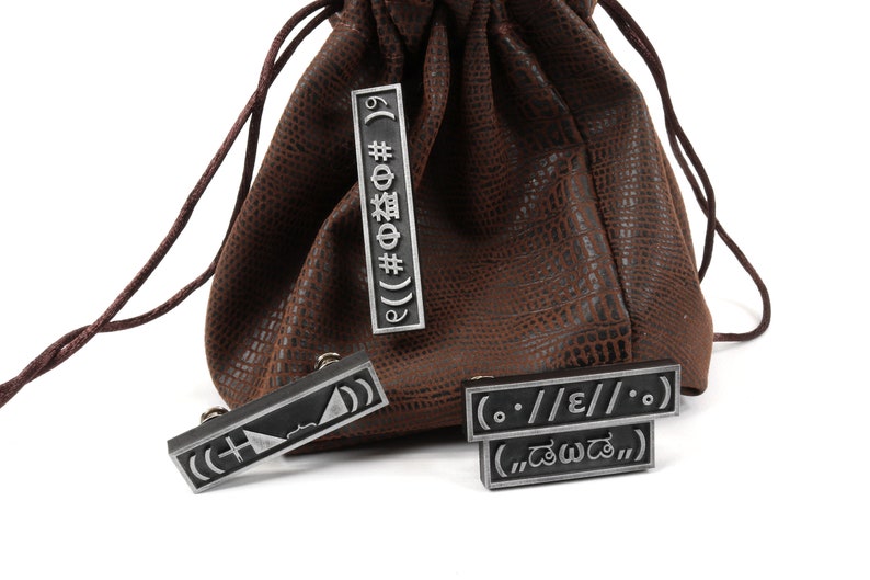 May include: A brown leather pouch with a drawstring closure and four silver metal tags with various symbols and text, including "((#Ф益# 6 &Phi;# 19)", "(..//E//..)", and "(.. డబ్బా ..)".