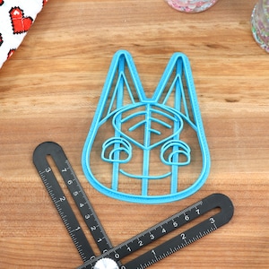 May include: A blue plastic cookie cutter shaped like the face of a cartoon animal with large ears and a smiling mouth.