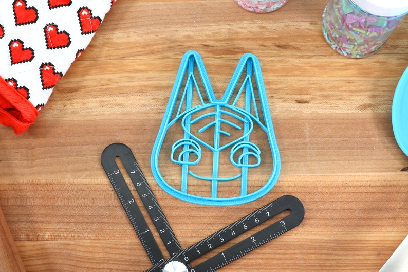 May include: A blue plastic cookie cutter shaped like the face of a cartoon animal with large ears and a smiling mouth.