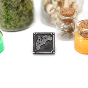 May include: A square, silver-colored metal charm with an ornate design, centered on a white surface. Small glass jars with cork stoppers and colorful contents, including green and orange liquids, are arranged around the charm.