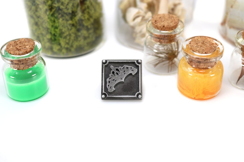 May include: A square, silver-colored metal charm with an ornate design, centered on a white surface. Small glass jars with cork stoppers and colorful contents, including green and orange liquids, are arranged around the charm.
