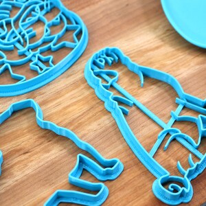 May include: A set of four blue plastic cookie cutters shaped like a person, a dog, a flower, and a star. The cutters are on a wooden surface.