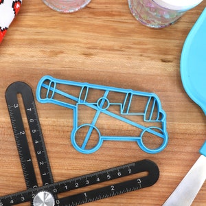 May include: A light blue plastic cannon-shaped cookie cutter sits on a wooden surface, next to a black angle ruler and a blue spatula. A white cloth with red pixelated hearts is in the background.