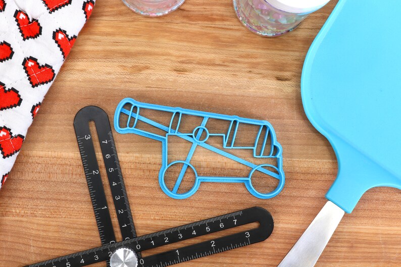 May include: A light blue plastic cannon-shaped cookie cutter sits on a wooden surface, next to a black angle ruler and a blue spatula. A white cloth with red pixelated hearts is in the background.