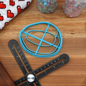 May include: A blue plastic cookie cutter shaped like a donut with a cross-hatch design. The cutter is on a wooden surface with a ruler, rolling pin, and a blue spatula.