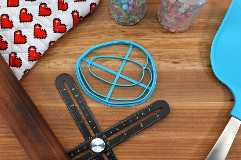 May include: A blue plastic cookie cutter shaped like a donut with a cross-hatch design. The cutter is on a wooden surface with a ruler, rolling pin, and a blue spatula.