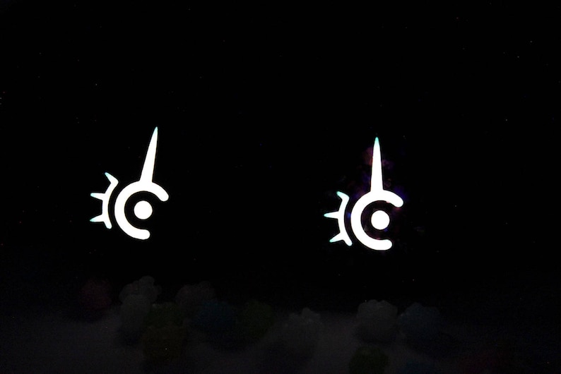 May include: Two white, stylized symbols with a circular base, a central dot, and a pointed top, set against a black background. The symbols have a sunburst-like design on the left side. The image has a glowing effect.